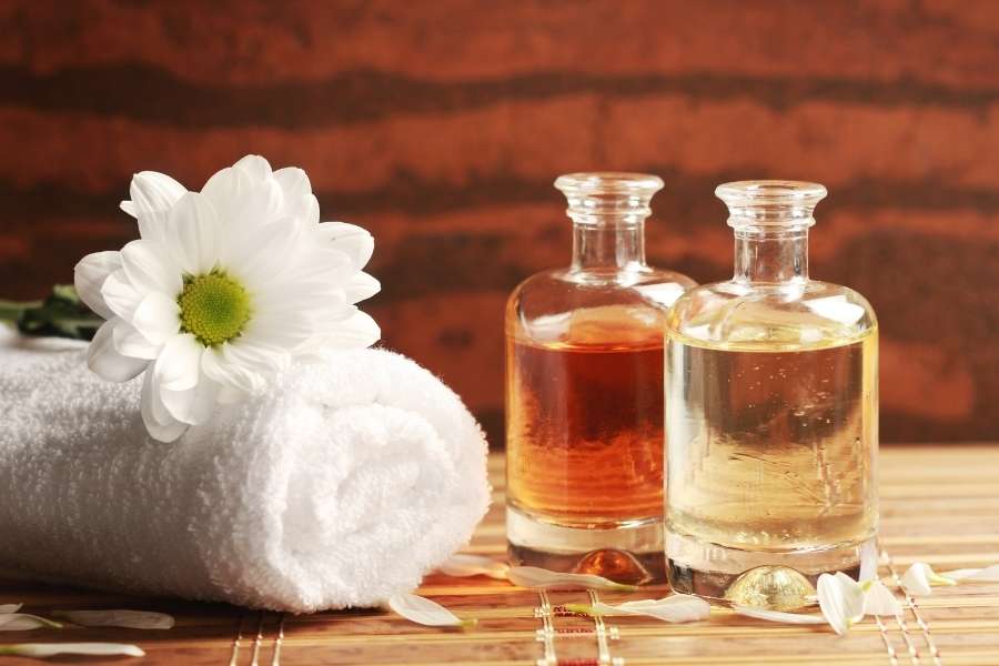 Deux flacons en verre remplis d'huiles de massage transparentes disposés à côté d'une serviette enroulée et d'une fleur blanche.