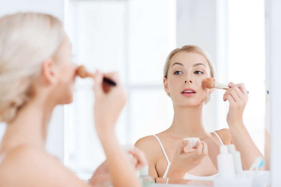 Une femme blonde se regarde dans un miroir lumineux et applique du blush avec un pinceau.