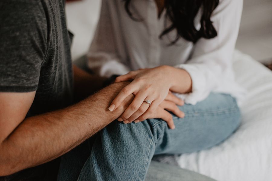 Un couple assis sur un lit, en jean et t-shirt/chemisier, où la main de l'homme tient celle de la femme, illustrant la communication des envies intimes.