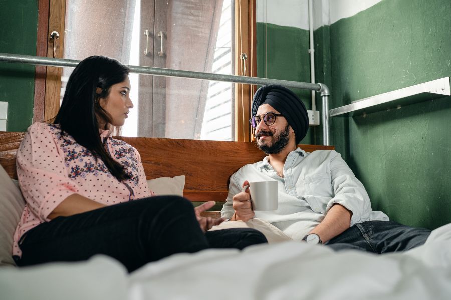 Un couple assis sur un lit, l'homme portant un turban, discute et se regarde intensément, symbolisant la communication des envies intimes.